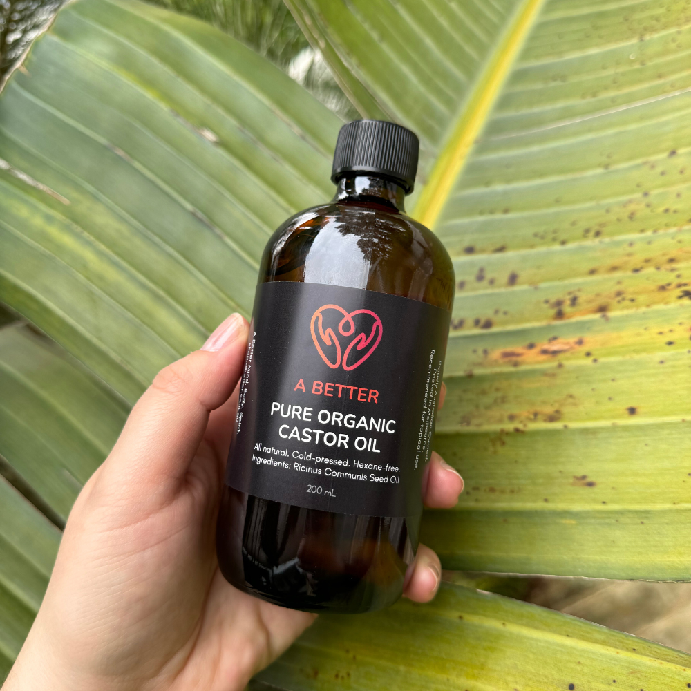 A hand holding a 200 mL amber glass bottle labeled 'A BETTER Pure Organic Castor Oil'.