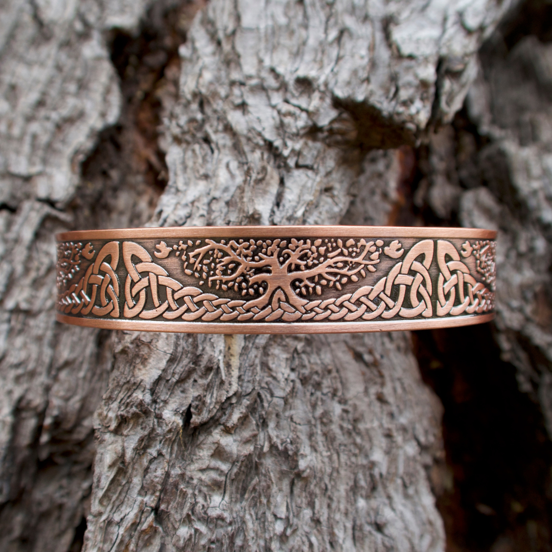 A copper cuff bracelet featuring an engraved Tree of Life pattern, shown against a close up of a tree trunk.