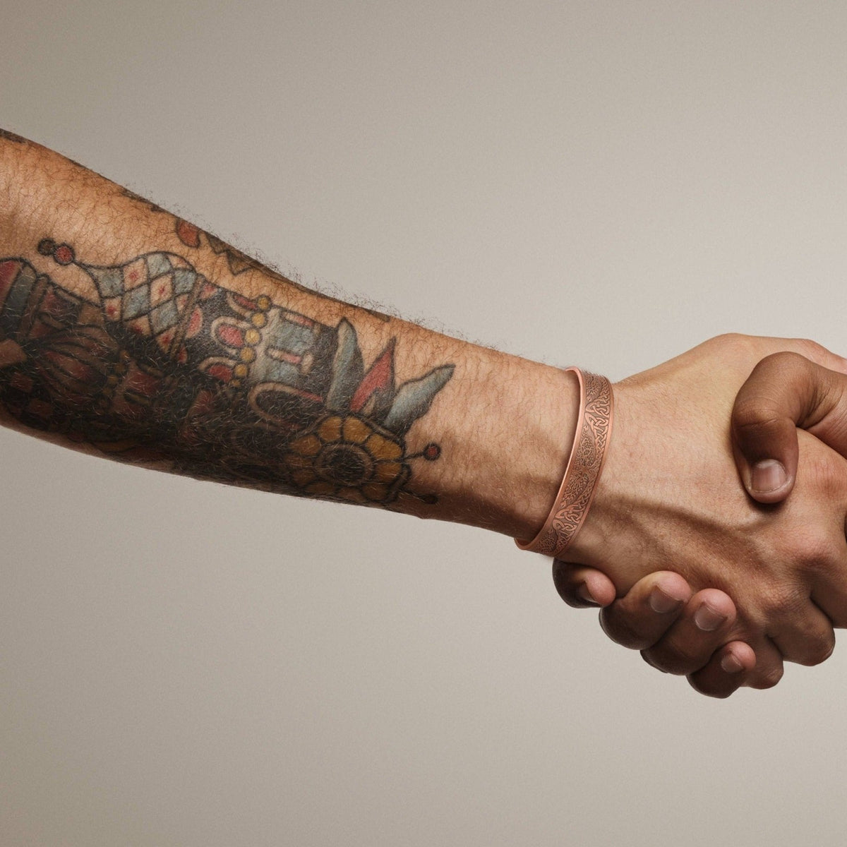 Two hands shaking hands, with one tattooed arm featuring a copper cuff bracelet, shown against a neutral background.
